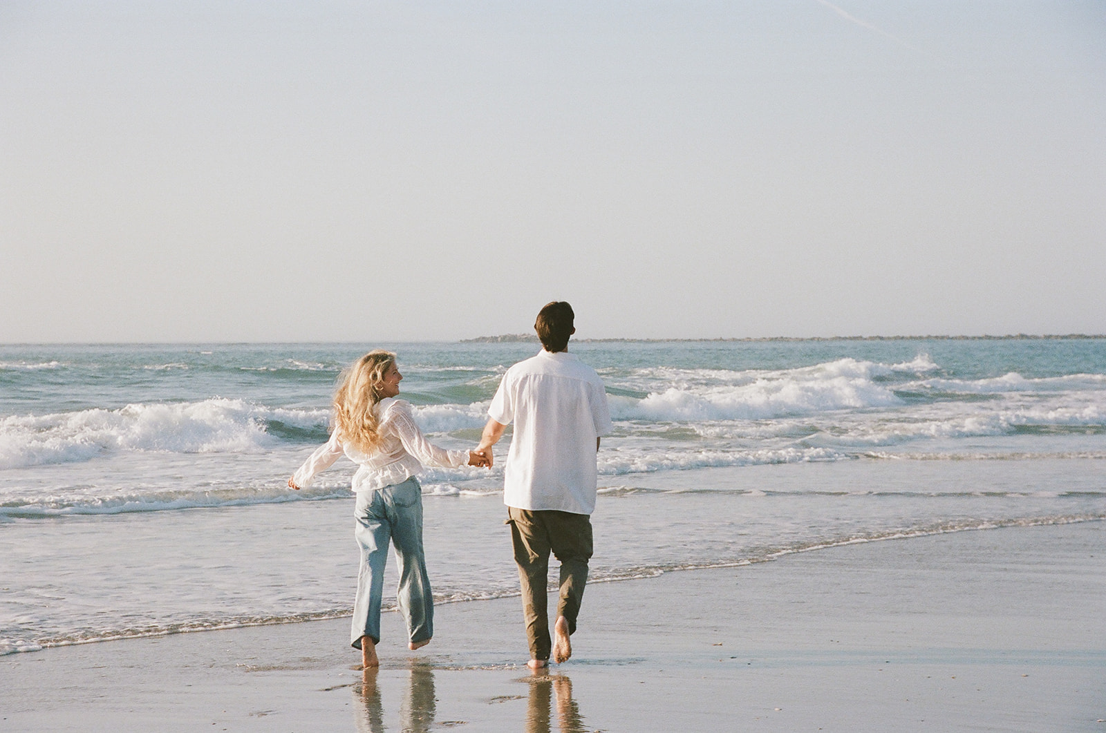 A person in a white top and blue jeans stands on a sandy beach near the shoreline with waves in the background.