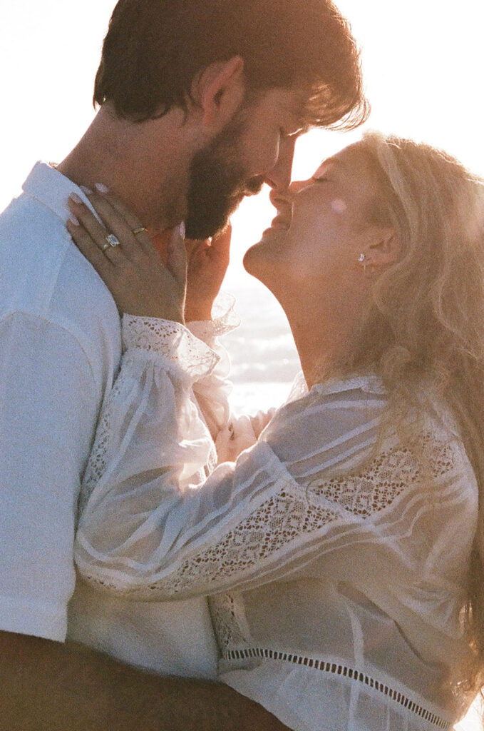 A couple wearing light-colored clothing embraces closely outdoors, with their faces touching and sunlight in the background.