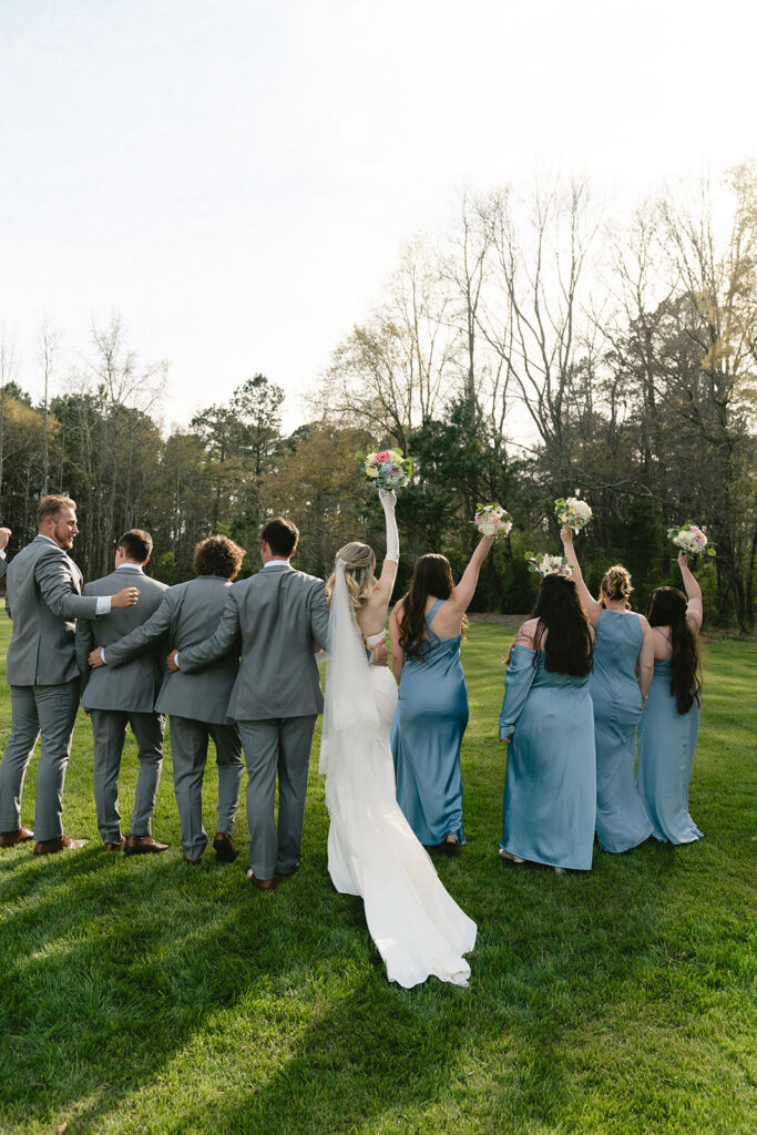 A bride, groom, groomsmen, and bridesmaids stand outdoors on grass, facing away, with the women raising bouquets in the air taken by a North Carolina wedding photographer