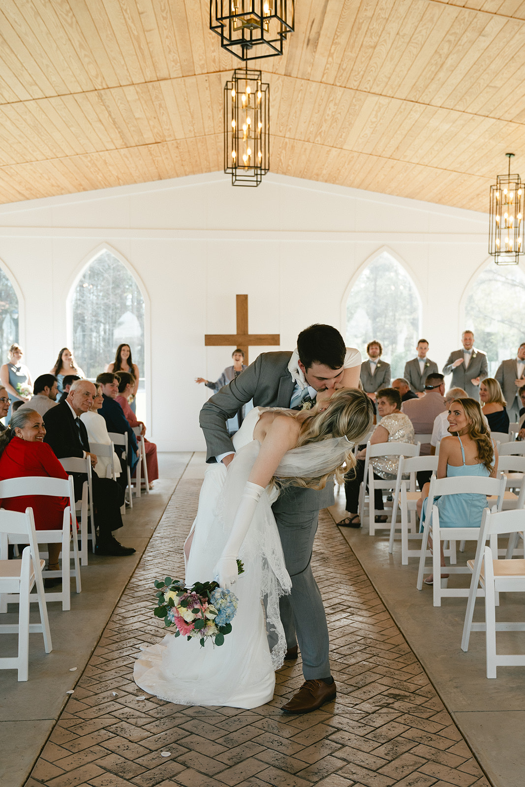 A bride and groom stand facing each other with an officiant in front of a large wooden cross during a wedding ceremony in a bright, white chapel taken by a North Carolina wedding photographer