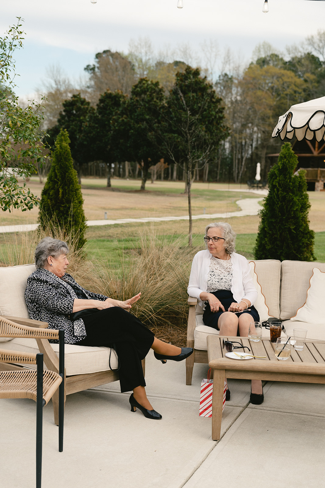 Five adults sit and talk around a small outdoor table under a white umbrella on a patio, with trees and a fireplace in the background.