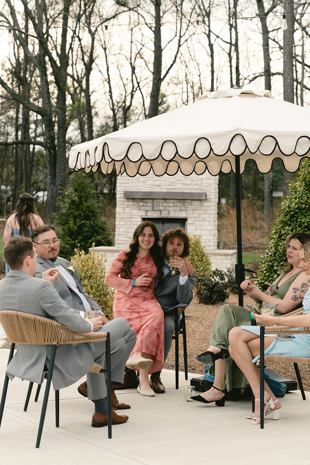Five adults sit and talk around a small outdoor table under a white umbrella on a patio, with trees and a fireplace in the background.