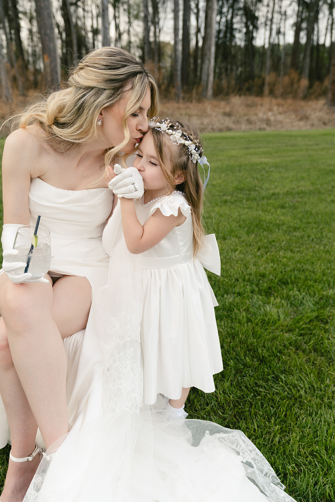 A bride in a white dress sits on grass, gently kissing the forehead of a young girl in a white dress and flower crown. Trees are visible in the background.