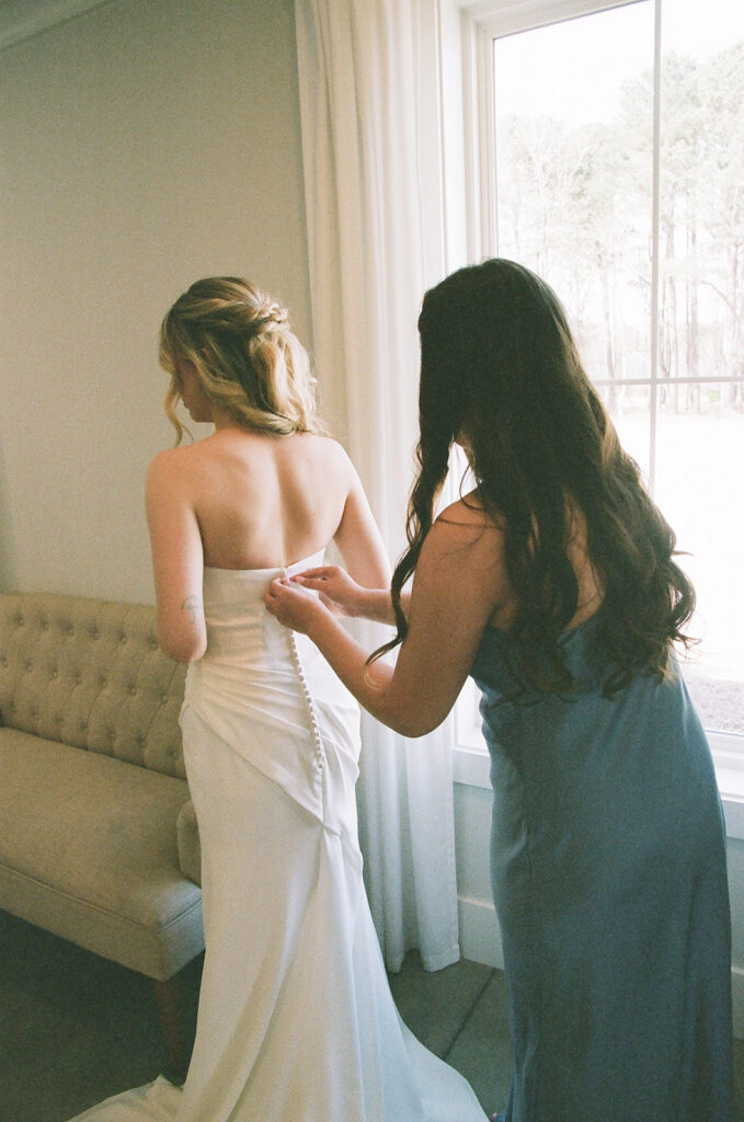 A woman in a light gown fastens the back of a bride’s white dress in a softly lit room near a window taken by a north carolina wedding photographer