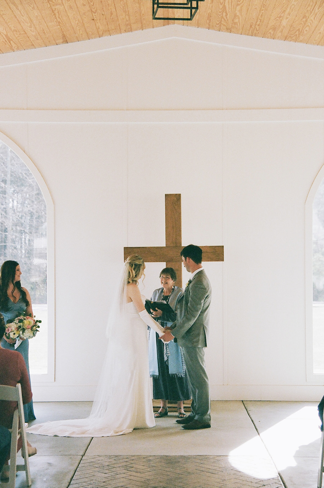 A bride and groom stand facing each other with an officiant in front of a large wooden cross during a wedding ceremony in a bright, white chapel taken by a North Carolina wedding photographer