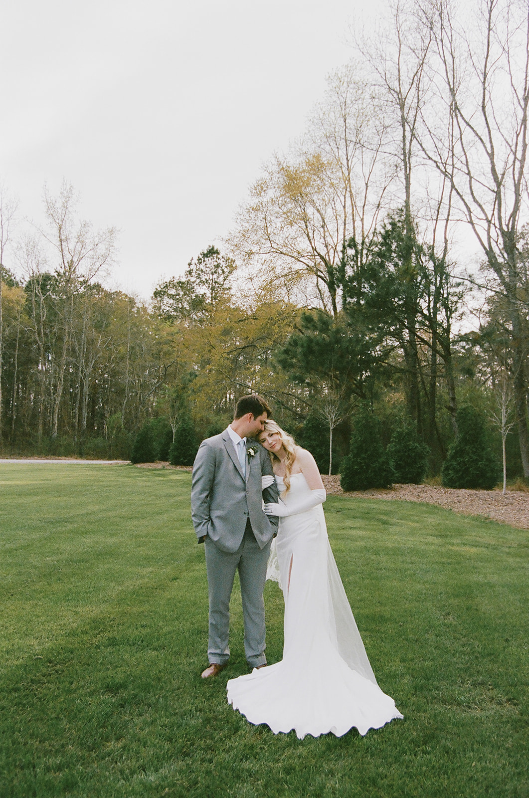 A bride and groom stand on grass, embracing each other closely with a wedding veil draped over both of them. Trees with early spring foliage are in the background.