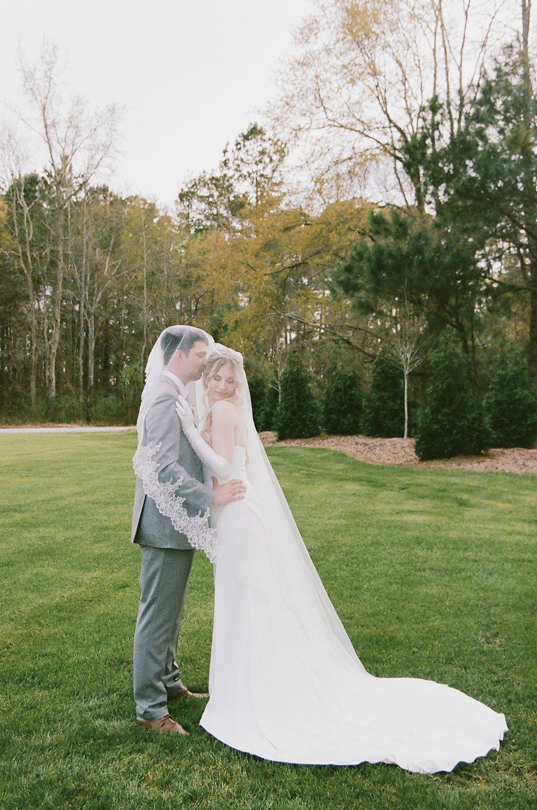 A bride and groom stand on grass, embracing each other closely with a wedding veil draped over both of them. Trees with early spring foliage are in the background.