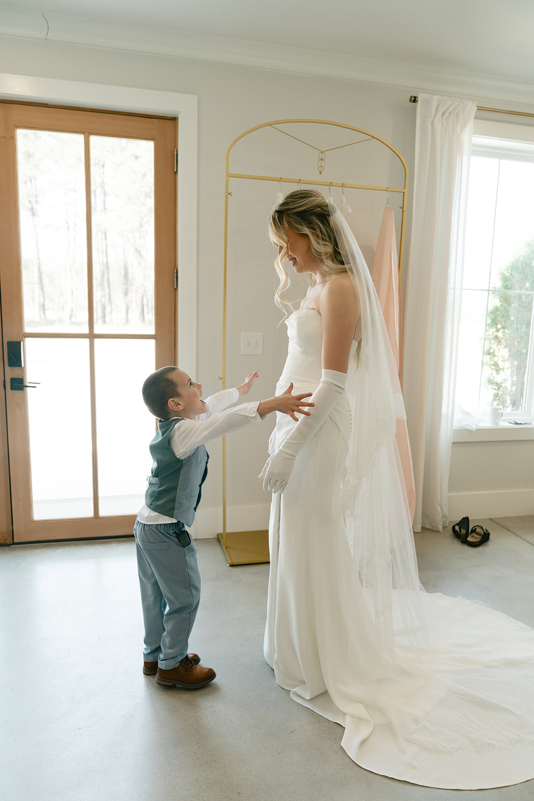 A bride in a white gown and veil stands facing a boy in a suit inside a bright room with large windows taken by a north carolina wedding photographer