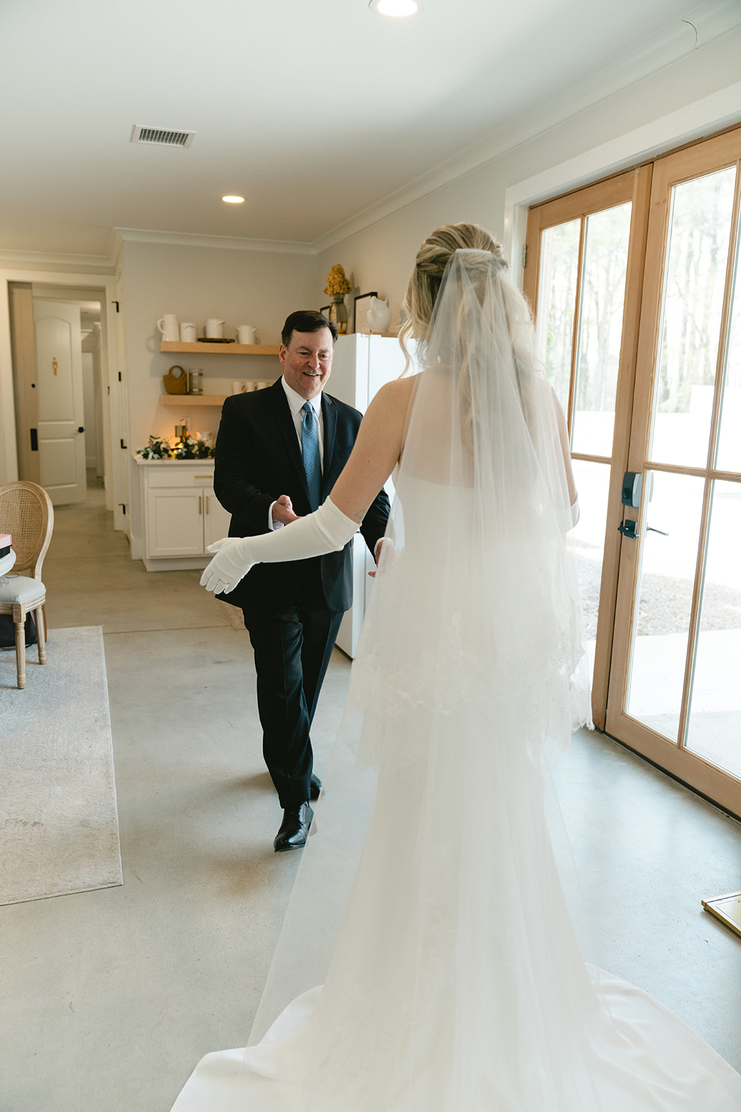 A bride in a white gown and veil stands facing a man in a suit inside a bright room with large windows taken by a north carolina wedding photographer
