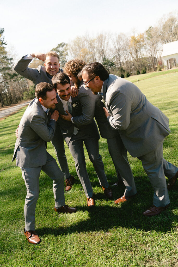 Five men in grey suits stand on grass, playfully huddling and laughing together on a sunny day with trees and a building in the background.