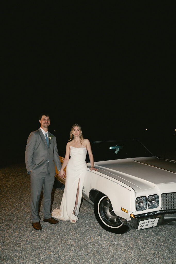 A man in a grey suit and a woman in a white dress stand next to a vintage white car at night on a gravel surface taken by a North Carolina wedding photographer