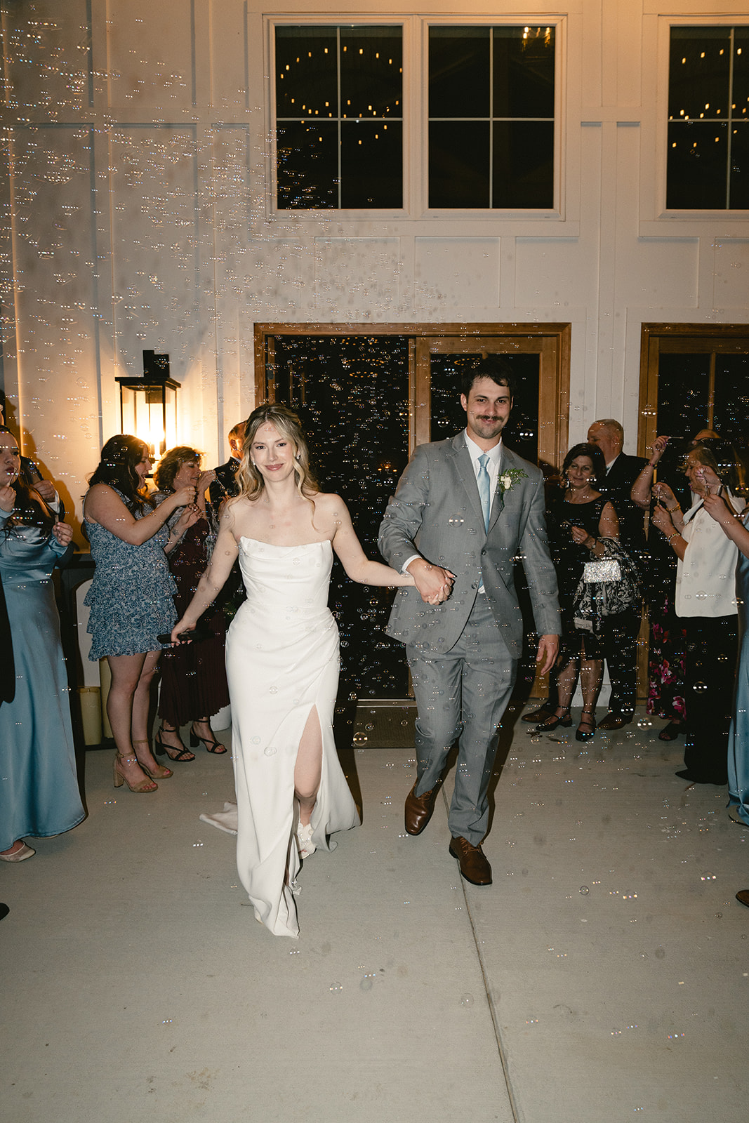 A bride and groom walk hand in hand through a crowd of guests indoors, with people cheering and bubbles in the air.