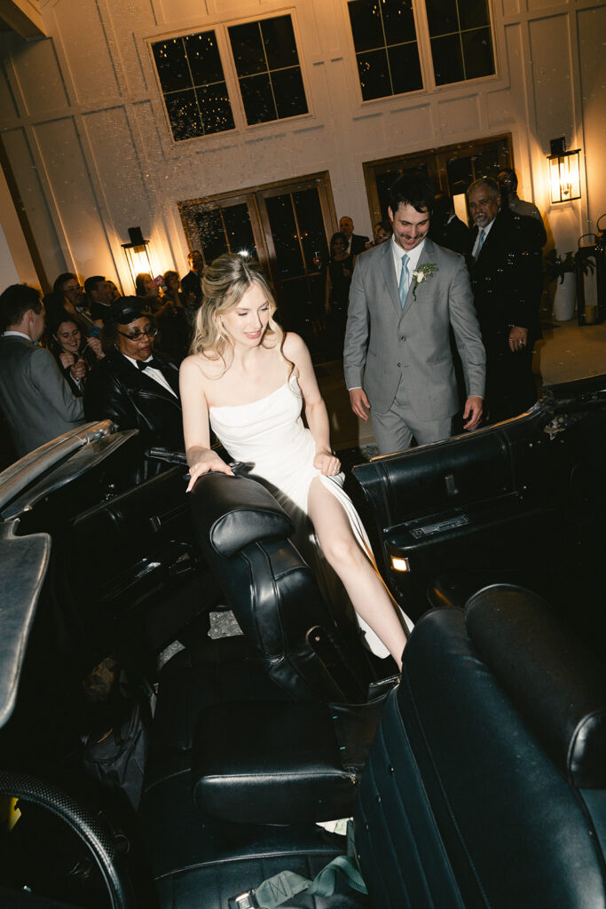 A woman in a white dress steps out of a black convertible at an indoor event, while a man in a suit follows behind her. Other guests are visible in the background.