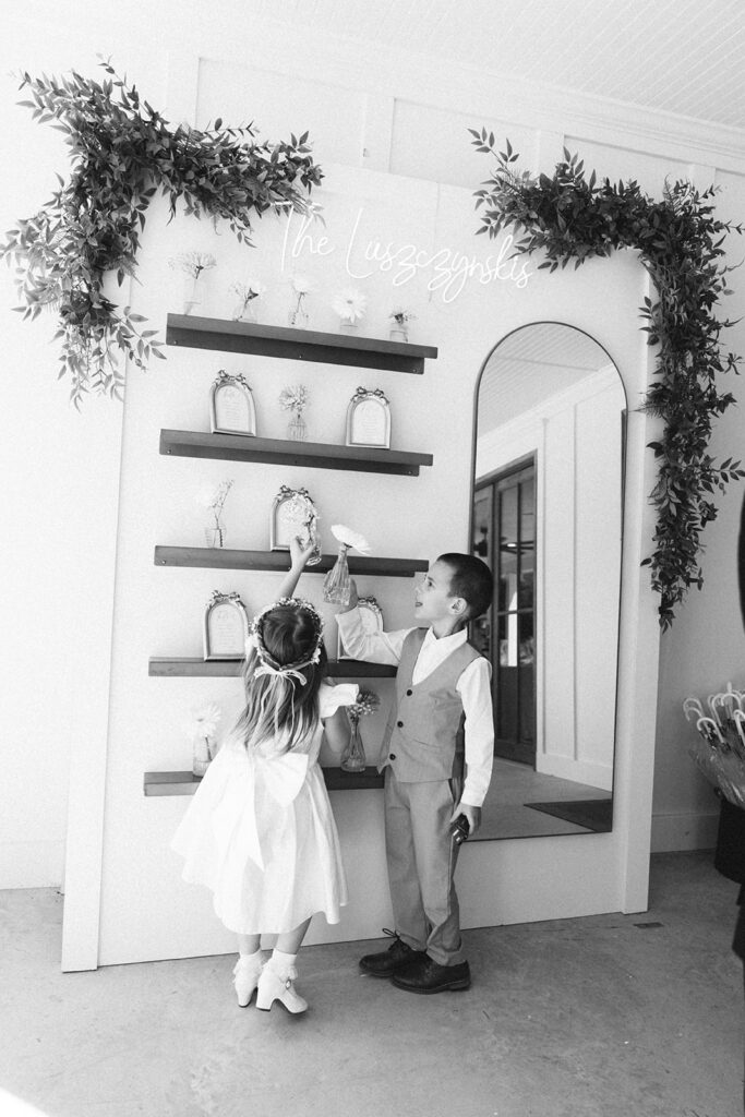 Two children dressed in formal attire stand in front of a decorated display shelf with small items and greenery.