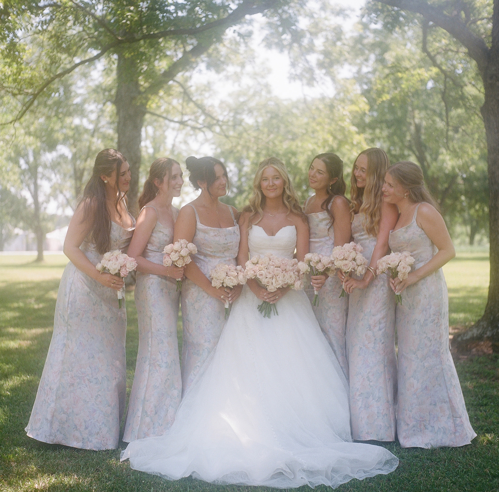 A bride and groom stand at the altar outdoors, surrounded by their wedding party and guests seated on wooden chairs under tall trees taken by a film wedding photographer