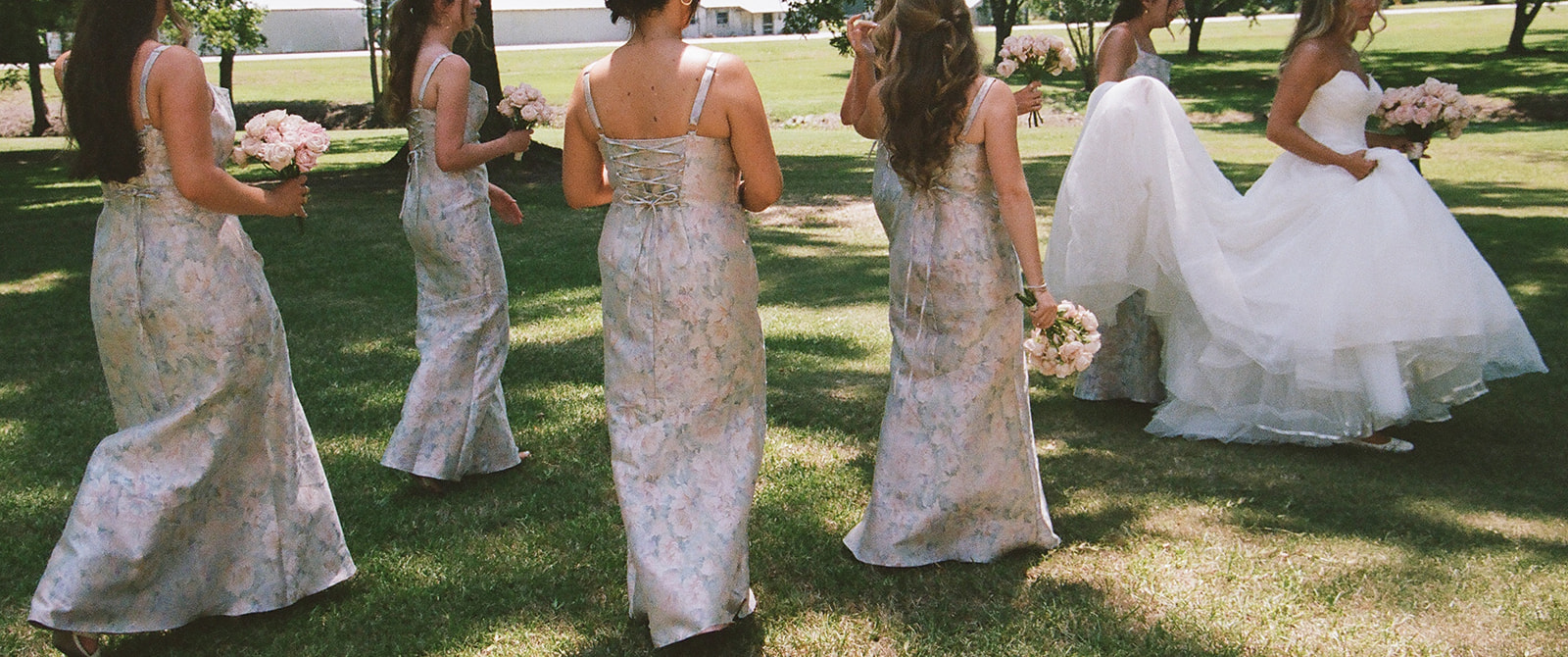 Six women in formal dresses, including one in a white wedding gown, walk together on a grassy lawn holding bouquets.