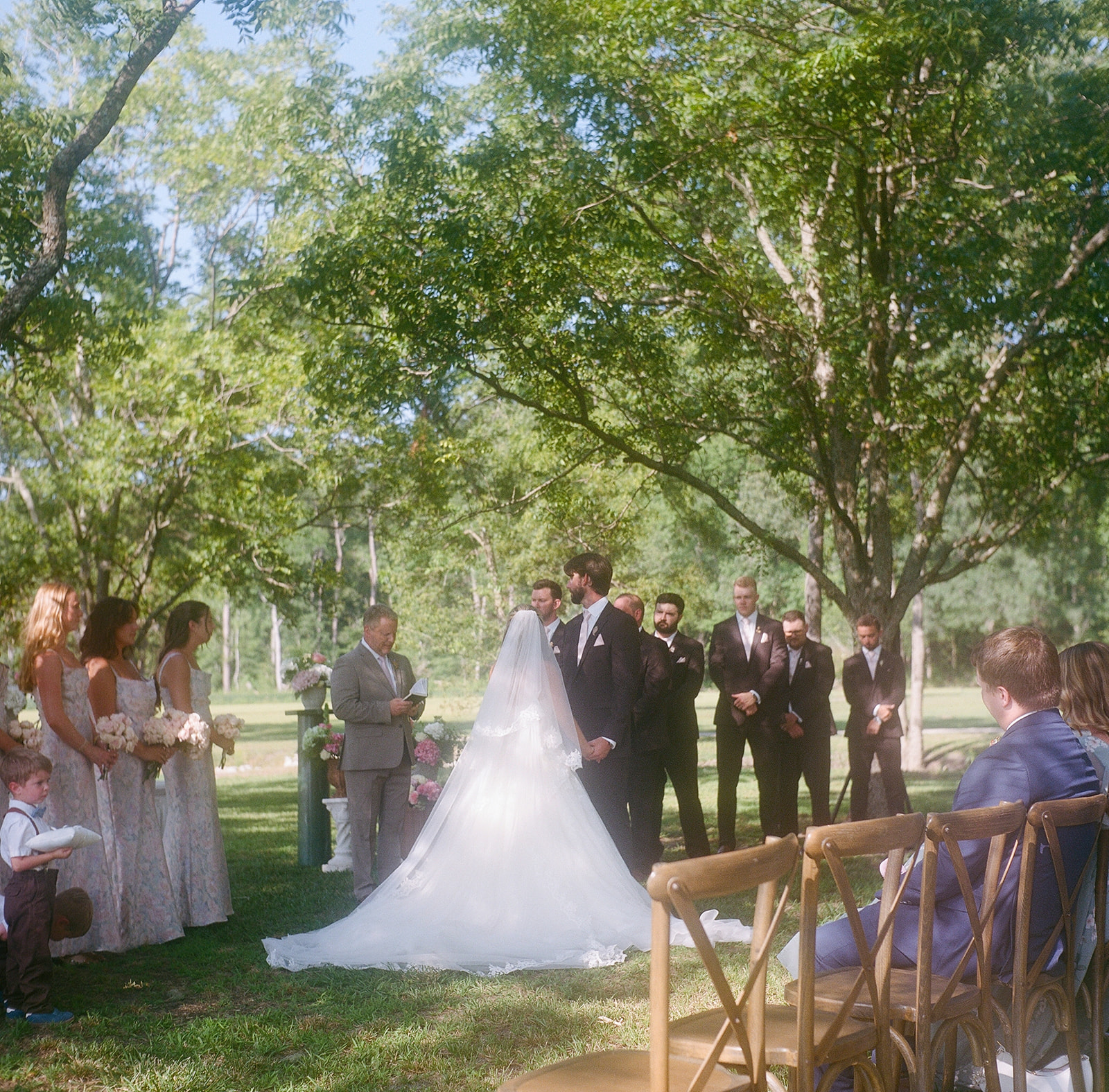 A bride and groom stand at the altar outdoors, surrounded by their wedding party and guests seated on wooden chairs under tall trees taken by a film wedding photographer