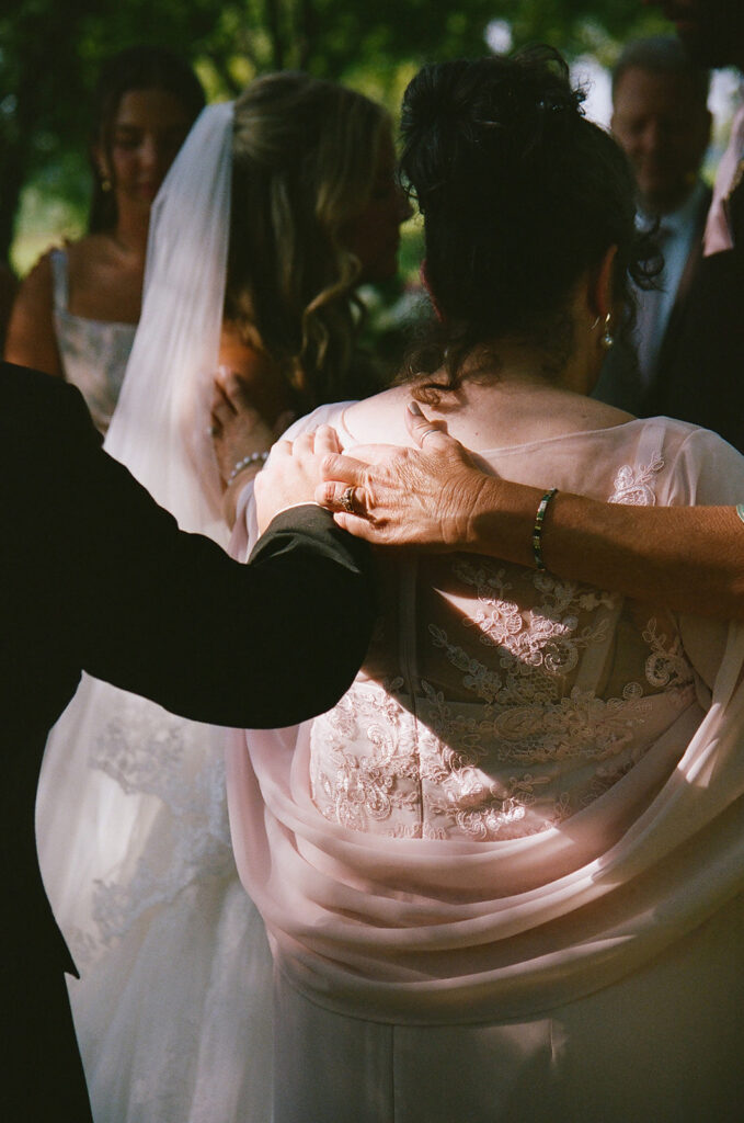 A group of people, including a bride in a veil, stand closely together outdoors, some with hands resting on each other’s backs taken by a film wedding photographer