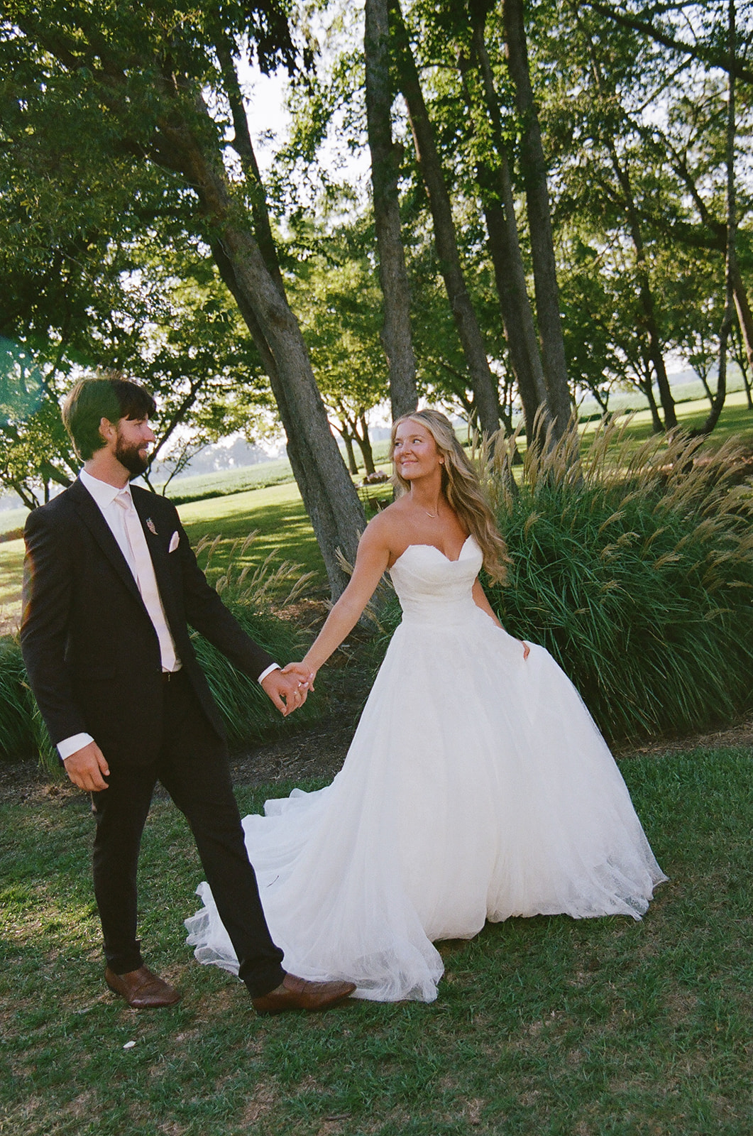 A bride in a white strapless gown and a groom in a dark suit hold hands and walk outdoors on grass, surrounded by green trees and sunlight.