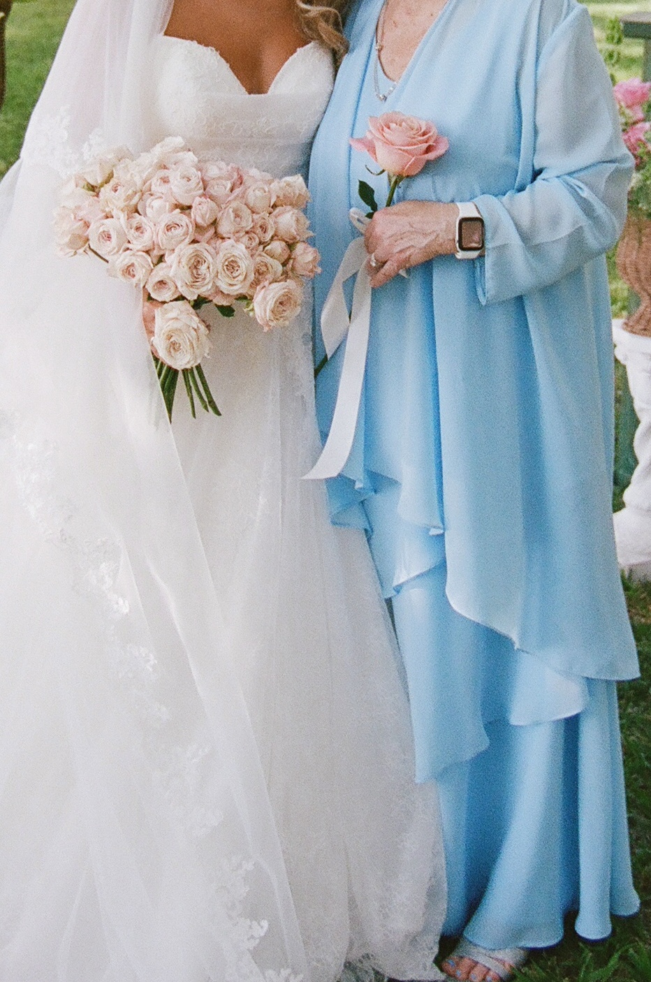 A bride in a white dress holding a bouquet stands beside a woman in a light blue dress holding a pink rose.