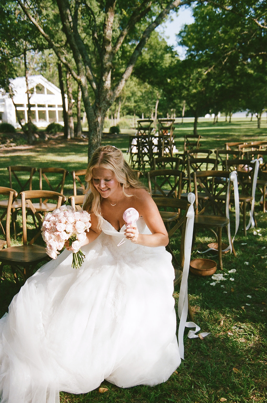 Bride in a white wedding dress sits on a wooden chair outdoors, holding a bouquet and eating ice cream, with empty chairs and trees in the background.