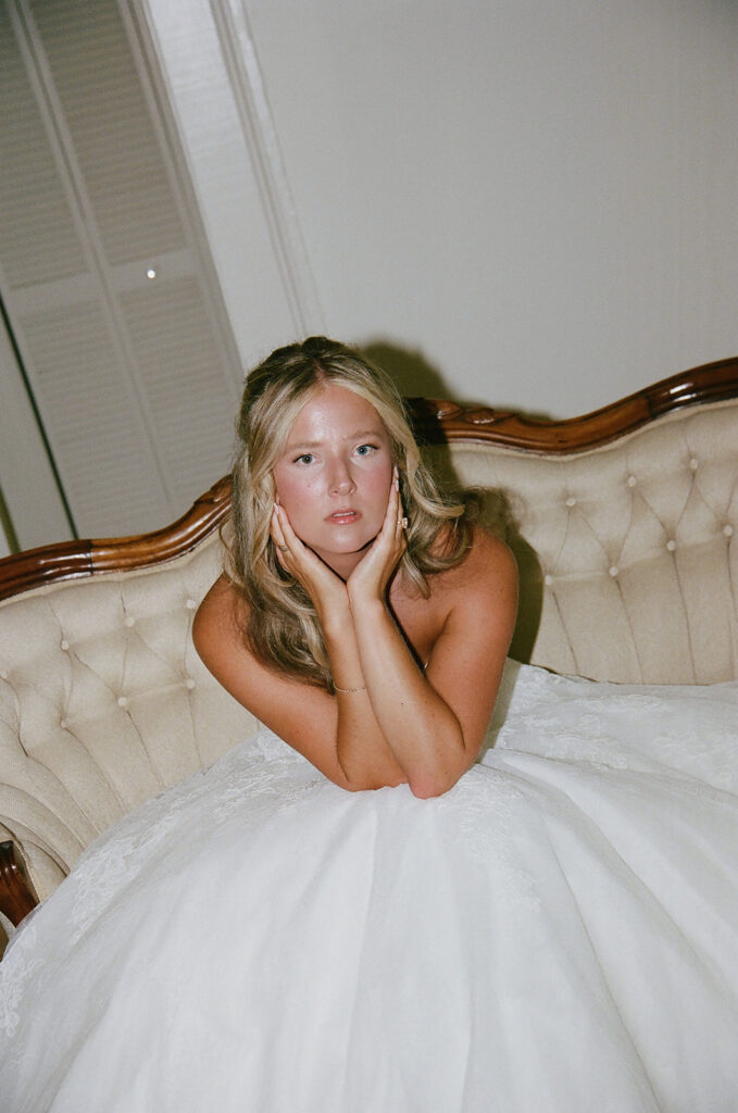 A woman in a white strapless dress sits on a cream tufted sofa, resting her chin on her hands and looking slightly upward.