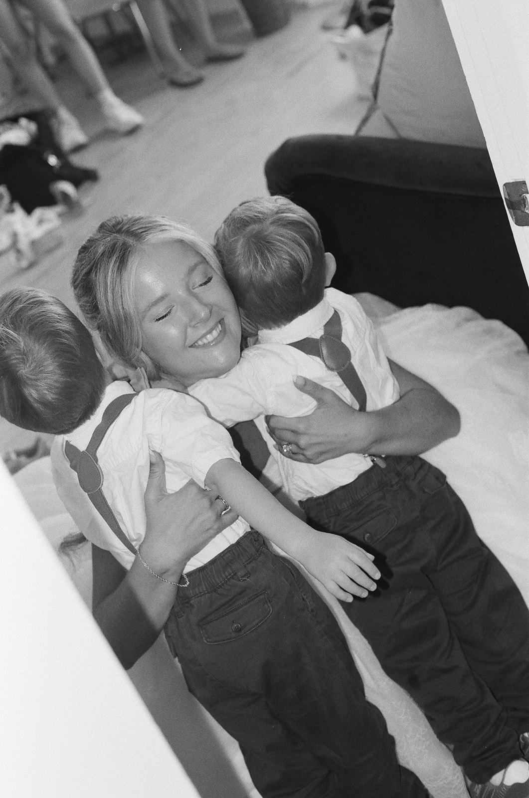 Woman smiling and hugging two young boys wearing matching outfits with suspenders in a candid indoor setting taken by a film wedding photographer