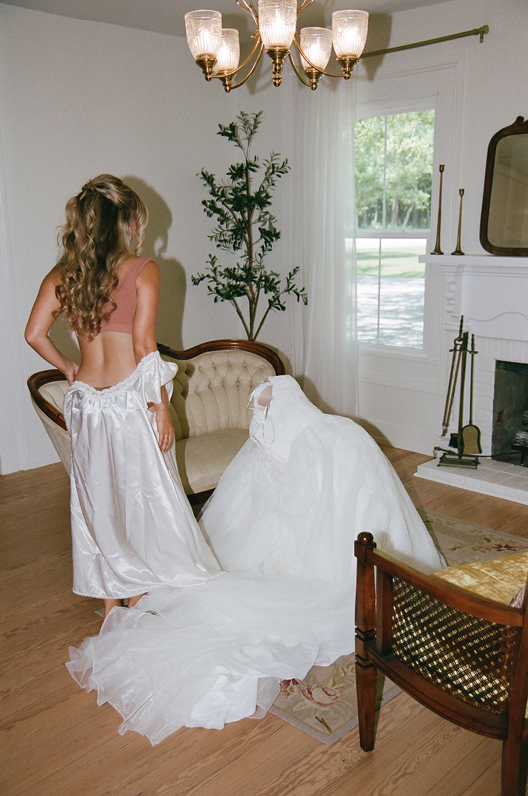 woman in wedding dresses stand in a living room; one faces away, revealing an open back, while the other sits on a cream sofa taken by a film wedding photographer