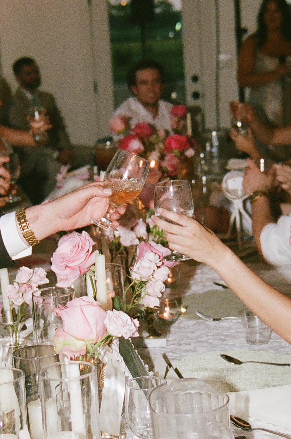 People seated at a table decorated with pink roses clink glasses in a toast during a gathering or celebration.