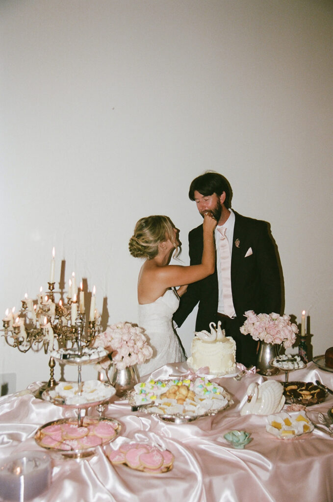 A bride in a white dress feeds cake to a groom in a suit beside a decorated dessert table with pink and white treats and candles taken by a film wedding photographer