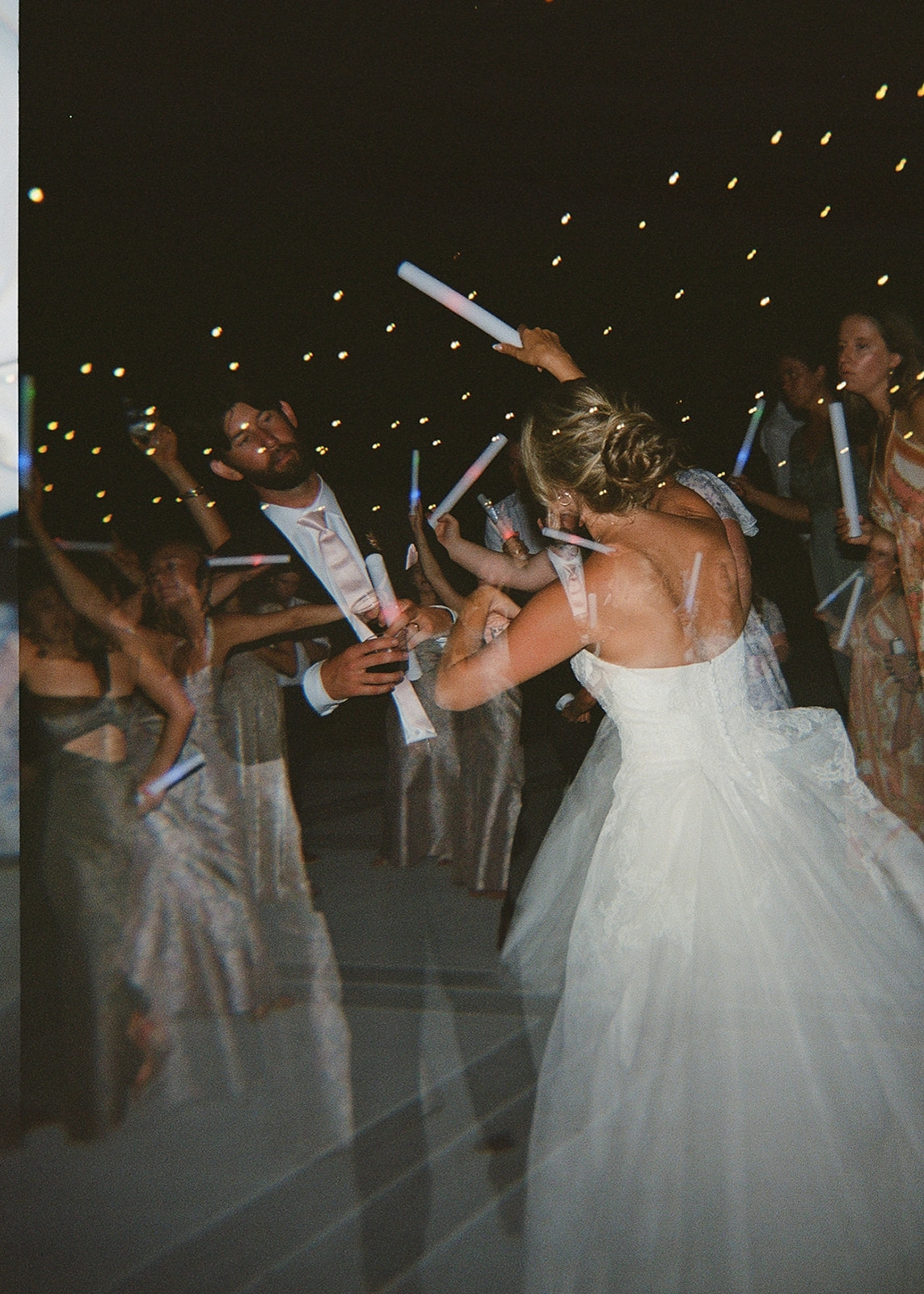 A bride and groom dance together at night, surrounded by guests holding glow sticks under string lights taken by a film wedding photographer