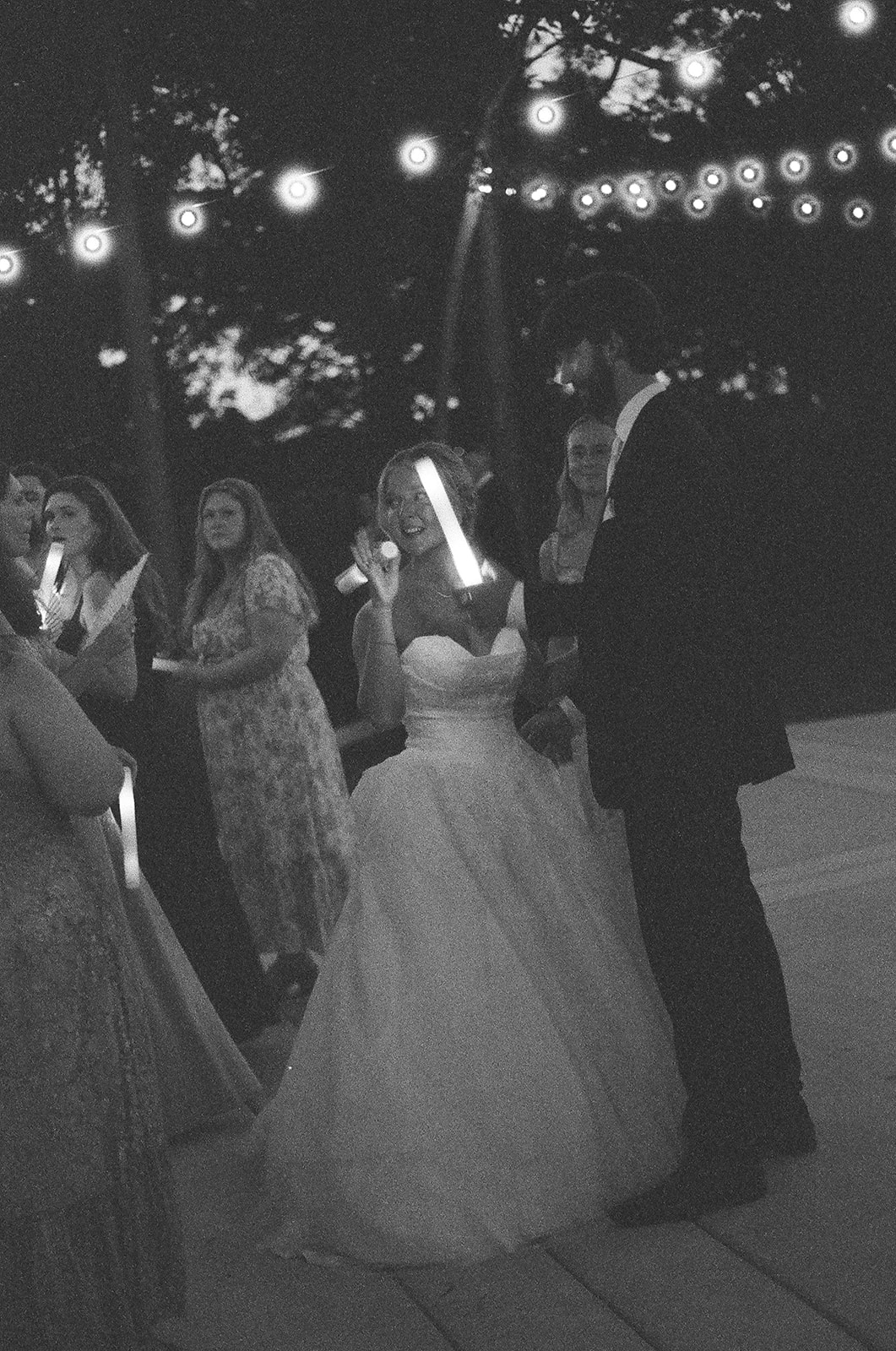 A bride and groom stand together outdoors at night, surrounded by guests under string lights.