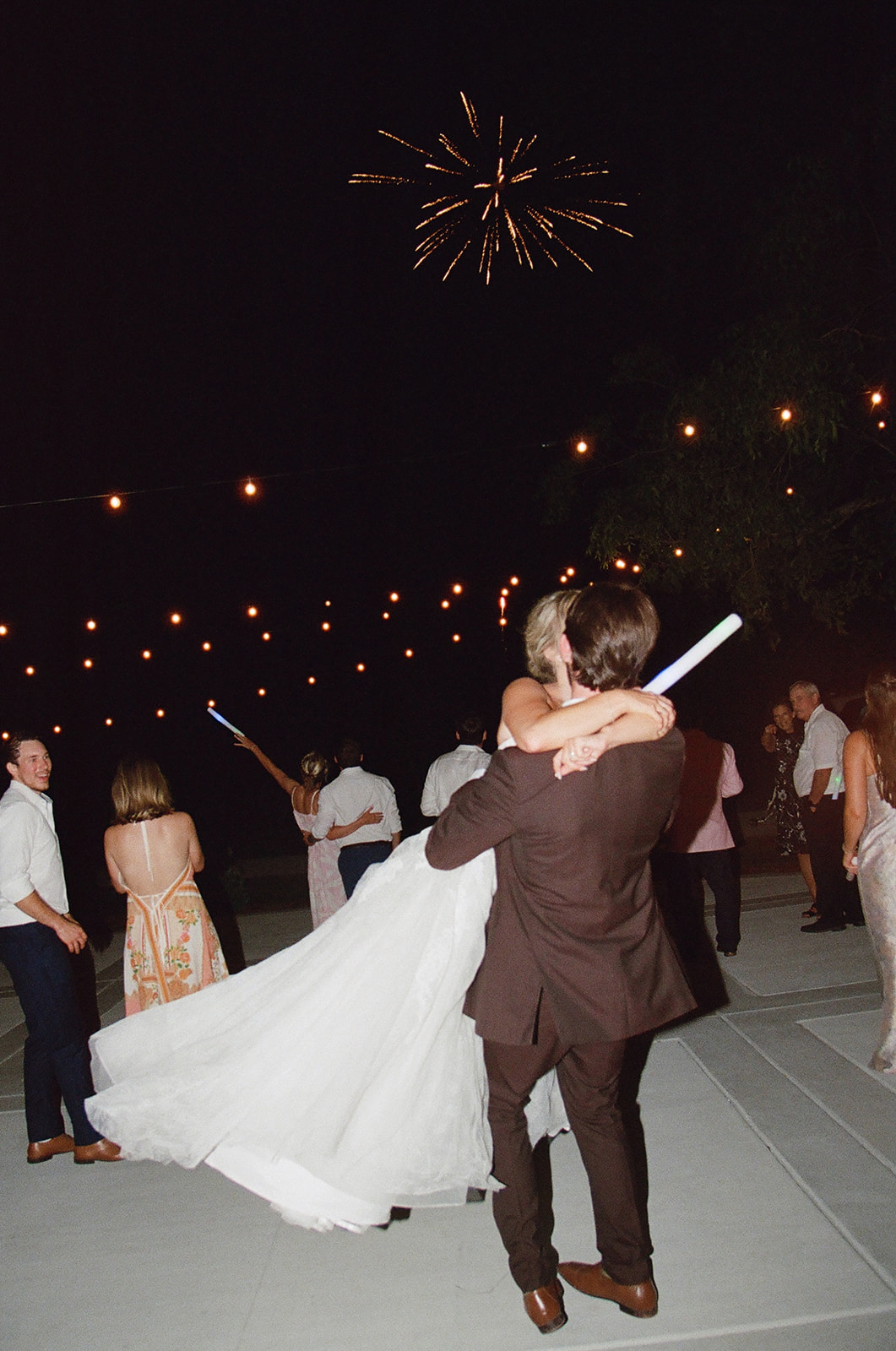A groom carries a bride in a white dress on a lit outdoor dance floor at night, with guests nearby and a firework exploding in the sky above.