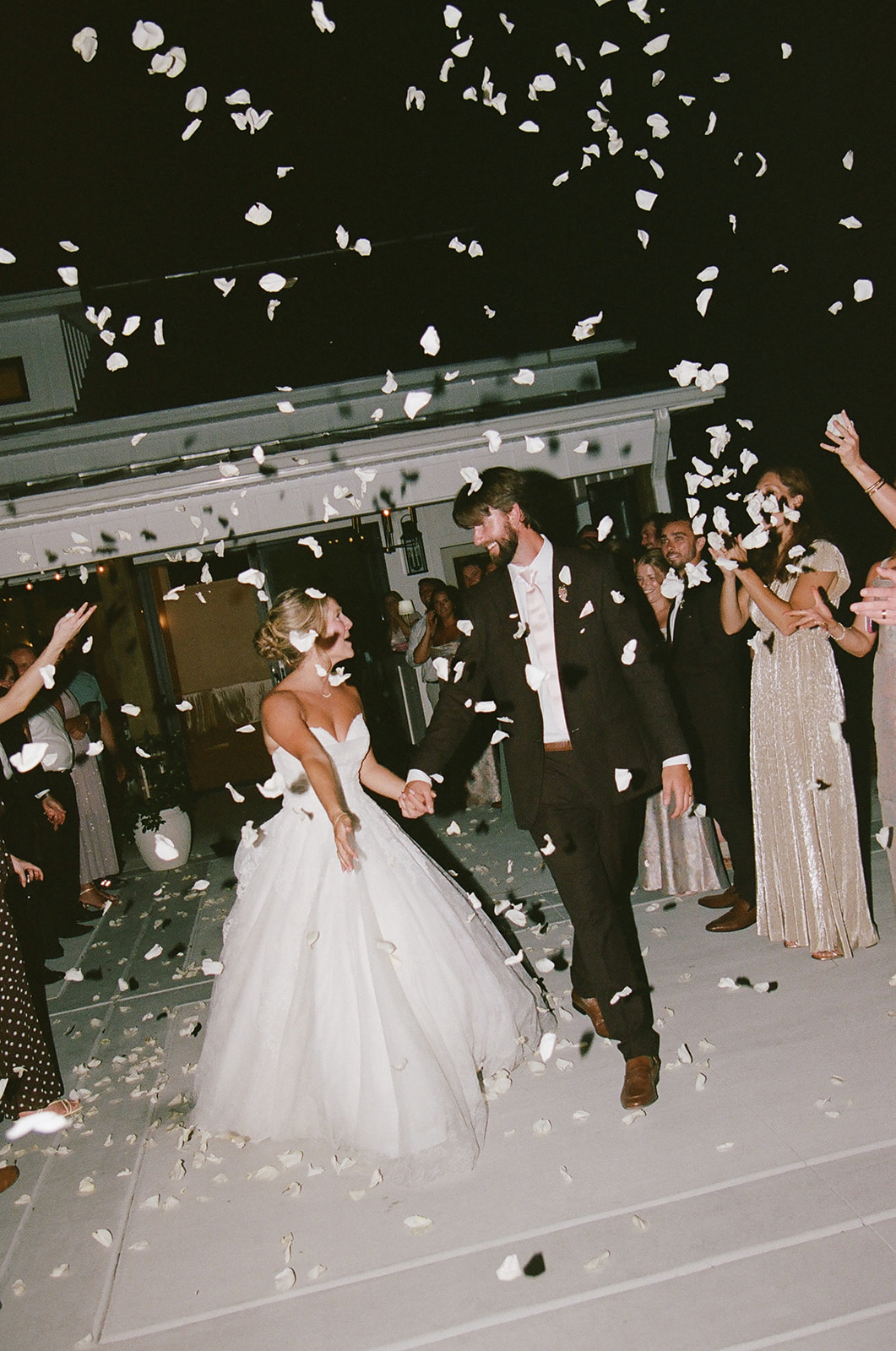 A bride and groom walk hand in hand while guests throw flower petals in the air during an outdoor nighttime wedding celebration.