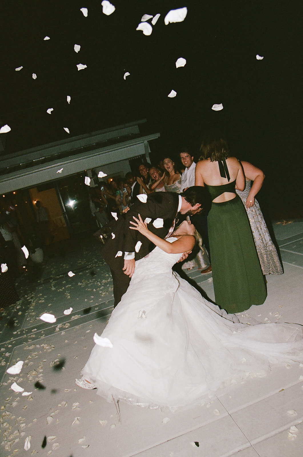 A bride and groom dance under falling flower petals at night, with guests standing and watching in the background taken by a film wedding photographer