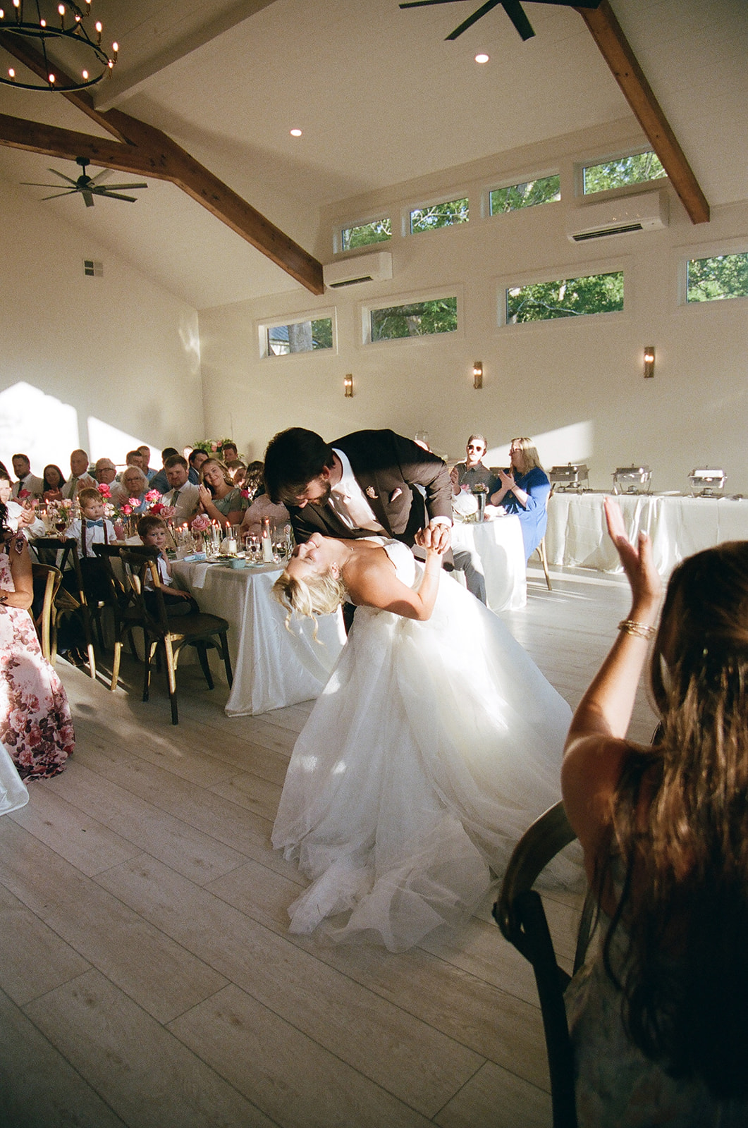 A bride and groom share a dip during their first dance at a wedding reception, surrounded by seated guests in a bright, modern venue.