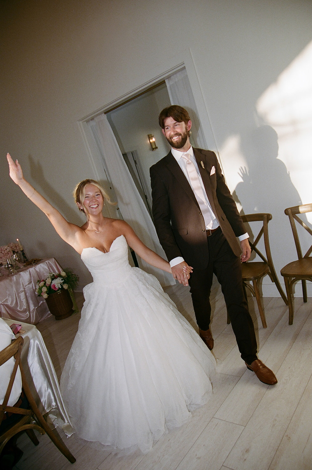 A bride in a white strapless gown and a groom in a black suit hold hands and smile as they enter a bright room with wooden chairs and floral decorations.